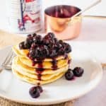 side shot of Blueberry sauce on pancakes staked on a plate with maple syrup