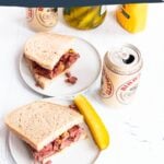 smoked meat sandwich on rye bread and mustard, a dill pickle on the side. Behind is a second half of a sandwich and two cans of beer.