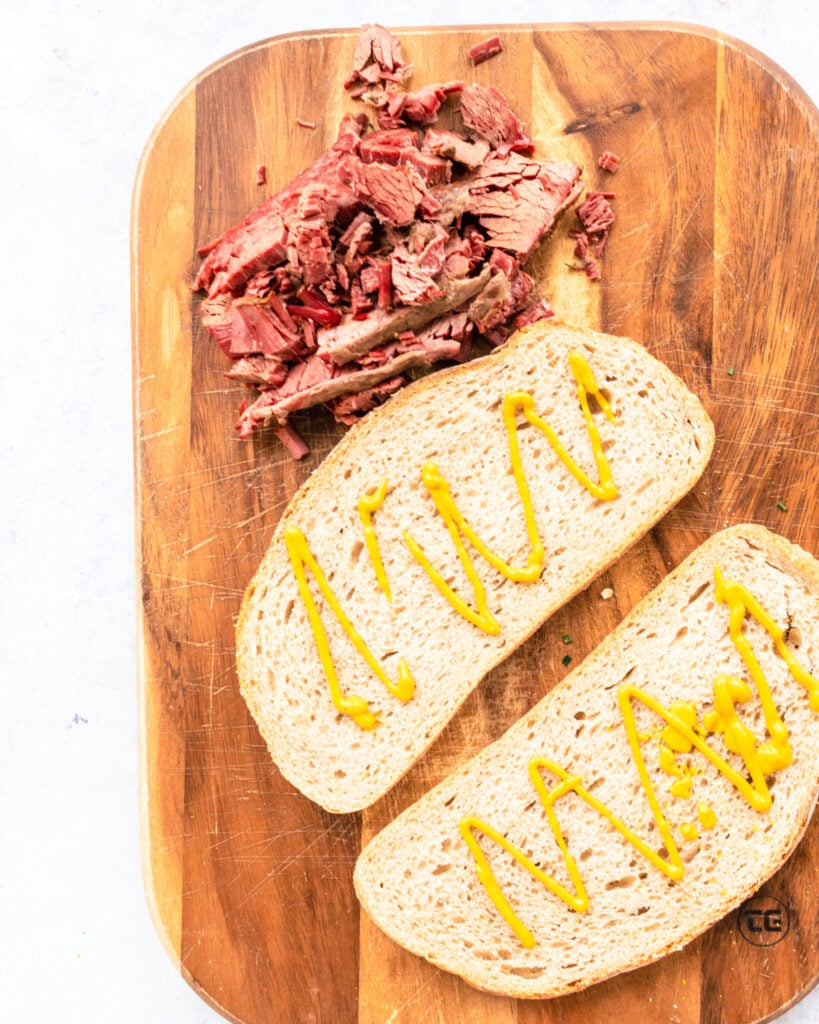 Two slices of rye bread with yellow mustard on them, a pile of chopped smoked meat is at the top of the photo