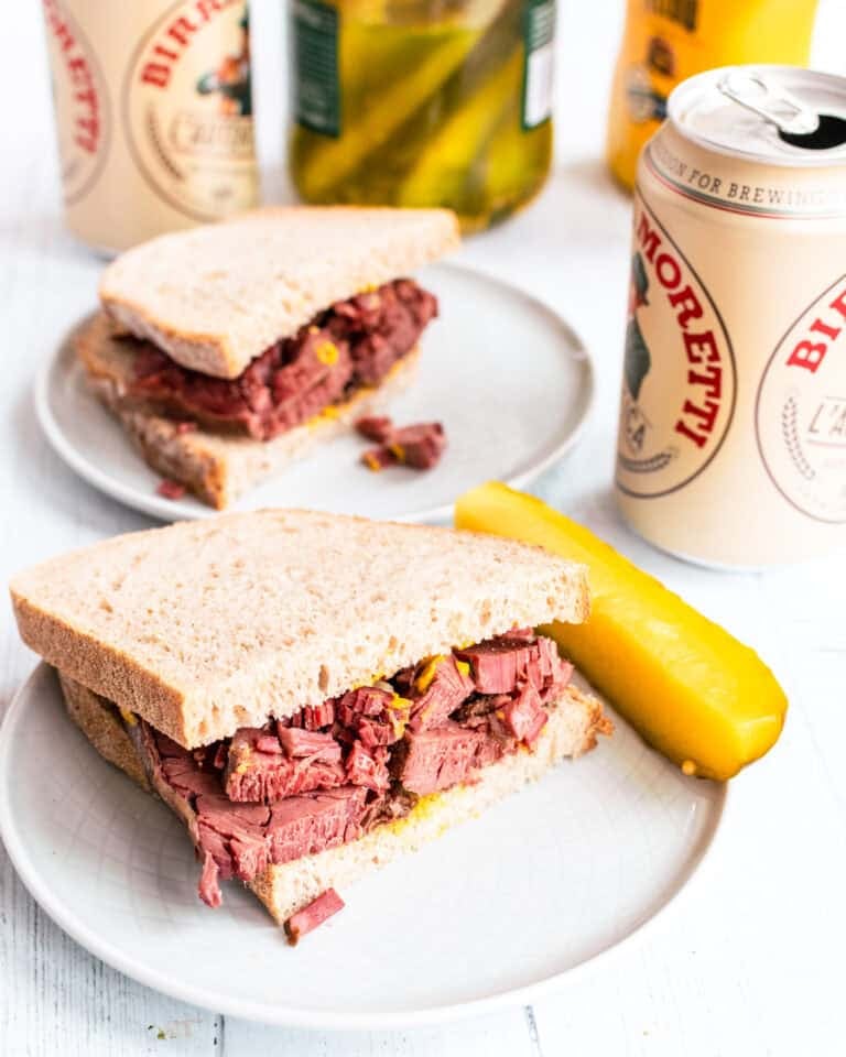 smoked meat sandwich on rye bread and mustard, a dill pickle on the side. Behind is a second half of a sandwich and two cans of beer.